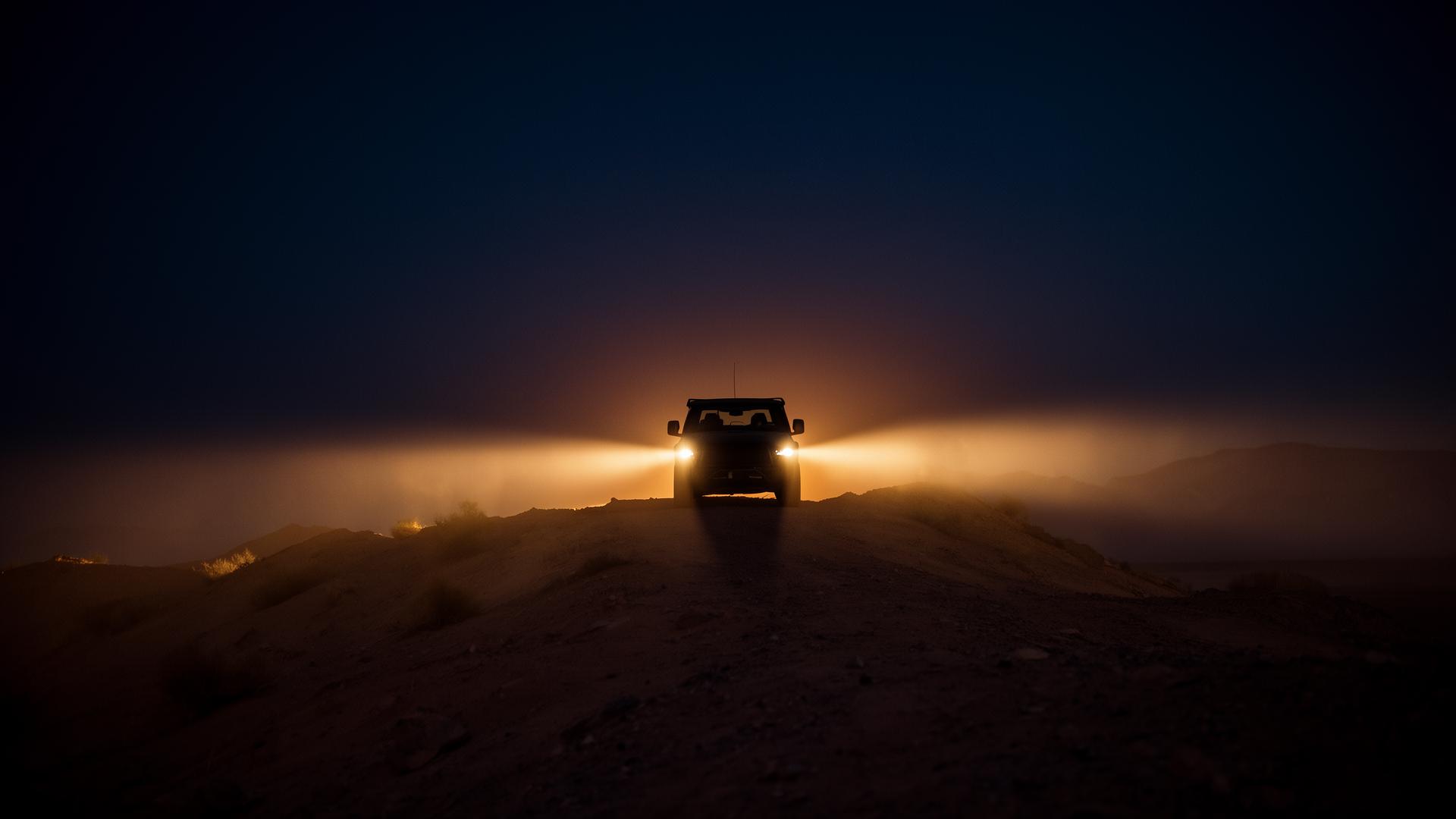 Cinematic dusk silhouette of an off-road vehicle on a desert ridge, headlight beams cutting through dust